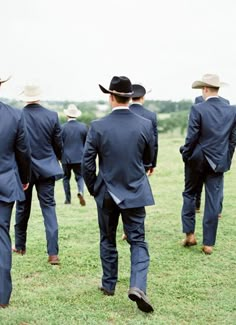 a group of men in suits and hats walking across a grass covered field with one man wearing a cowboy hat