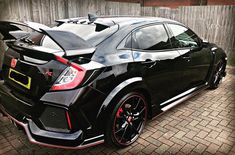 the rear end of a black sports car parked in front of a wooden fence on a brick road