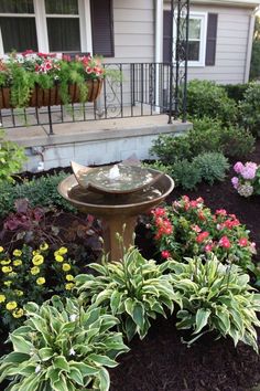 a bird bath in front of a house with flowers and plants around it's edges