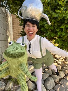 a woman is posing with a crocheted frog and a silver pot on her head