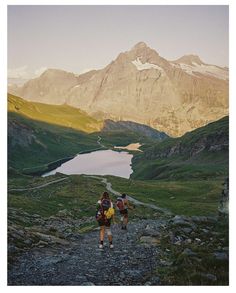 two people walking up a trail towards a lake in the mountains with snow capped peaks behind them