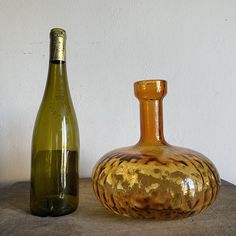 an empty wine bottle next to a large glass vase on a stone table with a white wall in the background