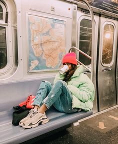 a woman sitting on a subway train with her face covered by a mask and wearing a red beanie