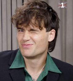 a man with curly hair wearing a green shirt and black blazer smiling at the camera