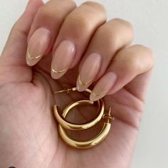 a woman's hand holding two gold hoop earrings and a pair of nude colored nails