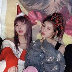 two young women sitting next to each other in front of a wall wearing christmas hats