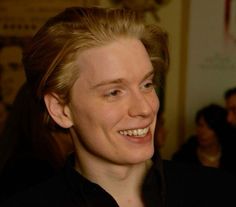 a woman with blonde hair smiling and wearing a black shirt in front of a group of people