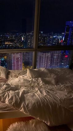 an unmade bed in front of a large window with city lights seen from it