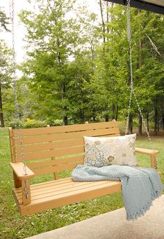 a porch swing with pillows on it and a blanket hanging from the back, in front of some trees