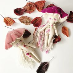 two cloth dolls sitting next to each other with leaves on the ground behind them and one doll wearing a pink hat