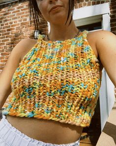 a woman with short hair wearing a colorful knitted crop top and white shorts standing in front of a brick building