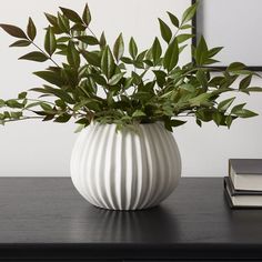 a white vase filled with green leaves on top of a wooden table next to a book