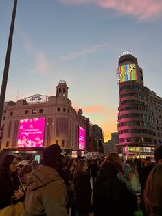 a crowd of people standing in the middle of a city at dusk with large buildings behind them