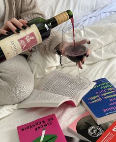 a person pouring wine into a glass next to several books and an open book on a bed