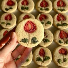 a hand holding a cookie decorated with red flowers and green leaves, in front of a tray of cookies