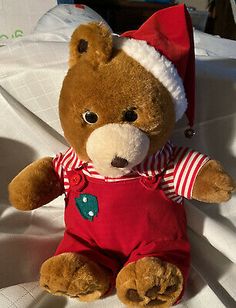 a brown teddy bear wearing a santa hat and red overalls sitting on a white sheet