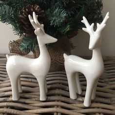 two white ceramic deer figurines sitting on a wicker table next to a pine tree