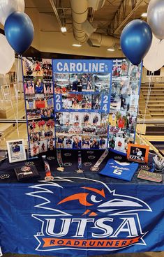 a table covered with pictures and balloons in the middle of a gym floor that is decorated with blue, white and orange banners