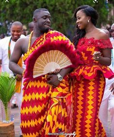 a man and woman dressed in traditional african garb, holding a fan while standing next to each other