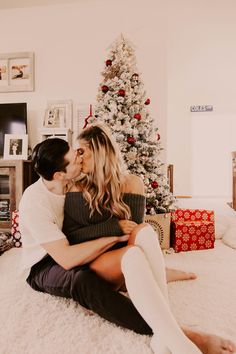 a man and woman sitting on the floor kissing in front of a christmas tree with presents