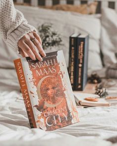 a person holding a book in their hand on top of a bed next to some books