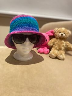 a white mannequin head wearing a pink and blue crocheted hat next to a brown teddy bear