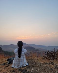 a woman sitting on top of a hill watching the sun go down in the distance