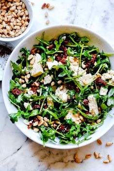 a white bowl filled with green salad next to another bowl full of nuts and cheese