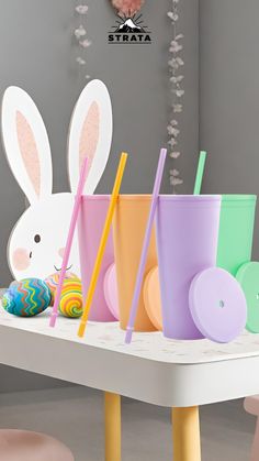 colorful cups with straws and bunny ears are on a table in front of an easter decoration