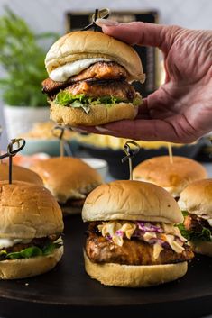 a hand holding a chicken sandwich with toothpicks in it and several other sandwiches on the table