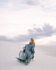 a woman standing in the sand with her dress blowing in the wind and clouds behind her