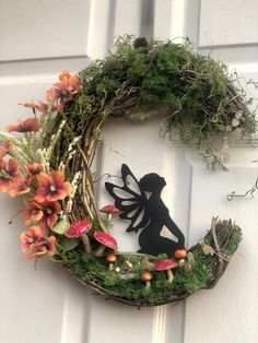 a wreath with a fairy on it and flowers hanging from the front door, next to a silhouette of a woman