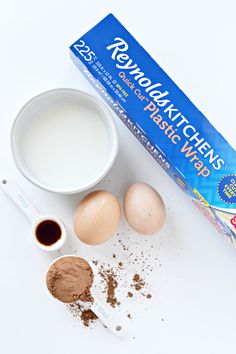 ingredients to make chocolate cake laid out on a white counter top, including eggs, milk, flour and baking utensils