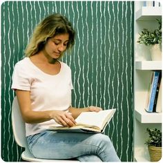 a woman sitting on a chair reading a book in front of a wall with bookshelves