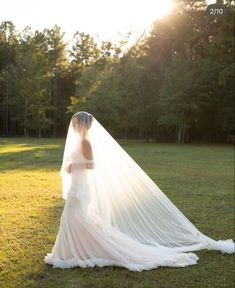 a woman in a wedding dress and veil standing on the grass with trees behind her