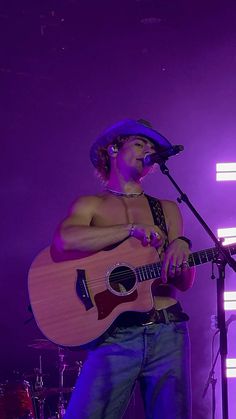 a man holding a guitar while standing in front of a microphone and wearing a cowboy hat