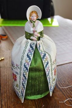 a small doll sitting on top of a green bottle covered in white fabric and ribbon