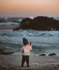 a little boy standing on top of a beach next to the ocean holding a cell phone