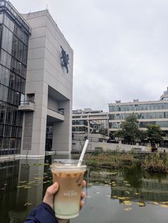 a person holding up a drink in front of a building with water and buildings around it