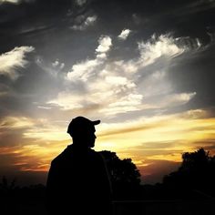 the silhouette of a man wearing a baseball cap and standing in front of a sunset