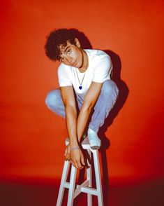 a young man sitting on top of a white stool in front of a red wall