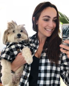 a woman holding a dog and taking a selfie with her cell phone while wearing a plaid shirt