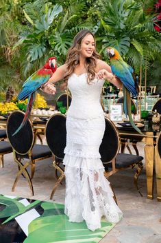 a woman in a white dress holding two parrots on her arm and posing for the camera