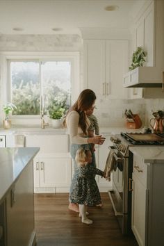 a woman holding a child while standing in a kitchen next to an open stove top oven