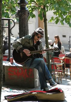 a man with a hat and sunglasses playing an acoustic guitar on the sidewalk in front of a cafe