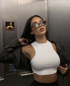 a woman in white top and black jacket standing next to metal wall with sunglasses on