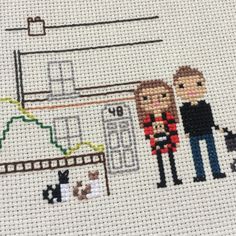 two people standing next to each other in front of a cross stitch pattern