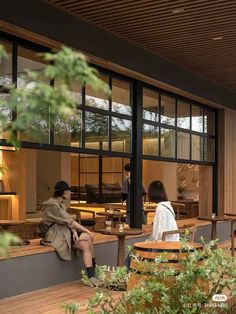 two people sitting on benches in front of a building with large windows and wood flooring