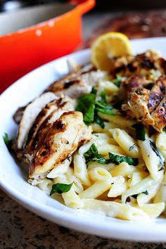 a white bowl filled with pasta and chicken on top of a wooden table next to an orange juice