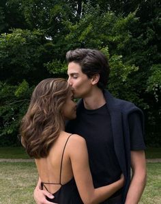 a young man and woman standing next to each other in front of green grass with trees behind them